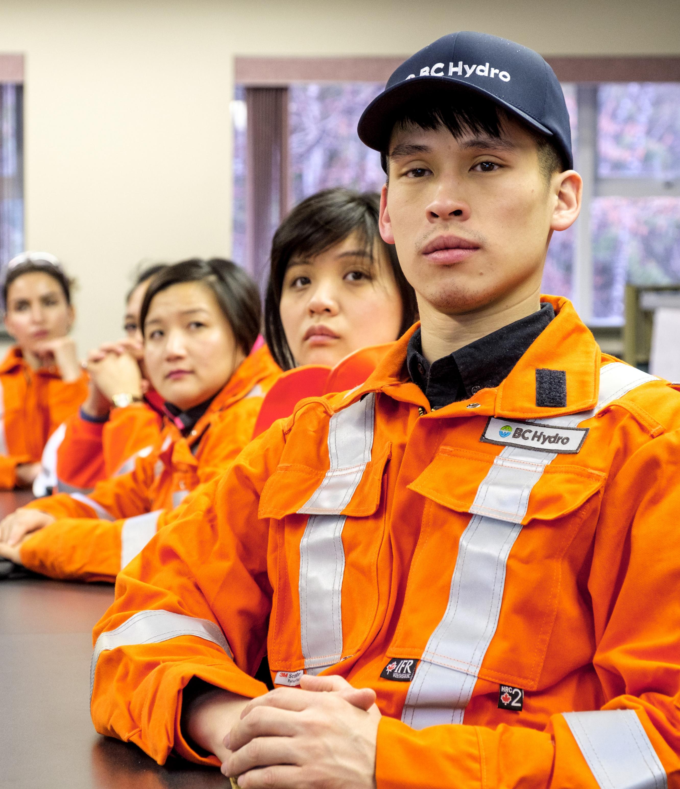 Several BC Hydro employees wearing orange safety jackets, sitting in a room together and facing the camera.