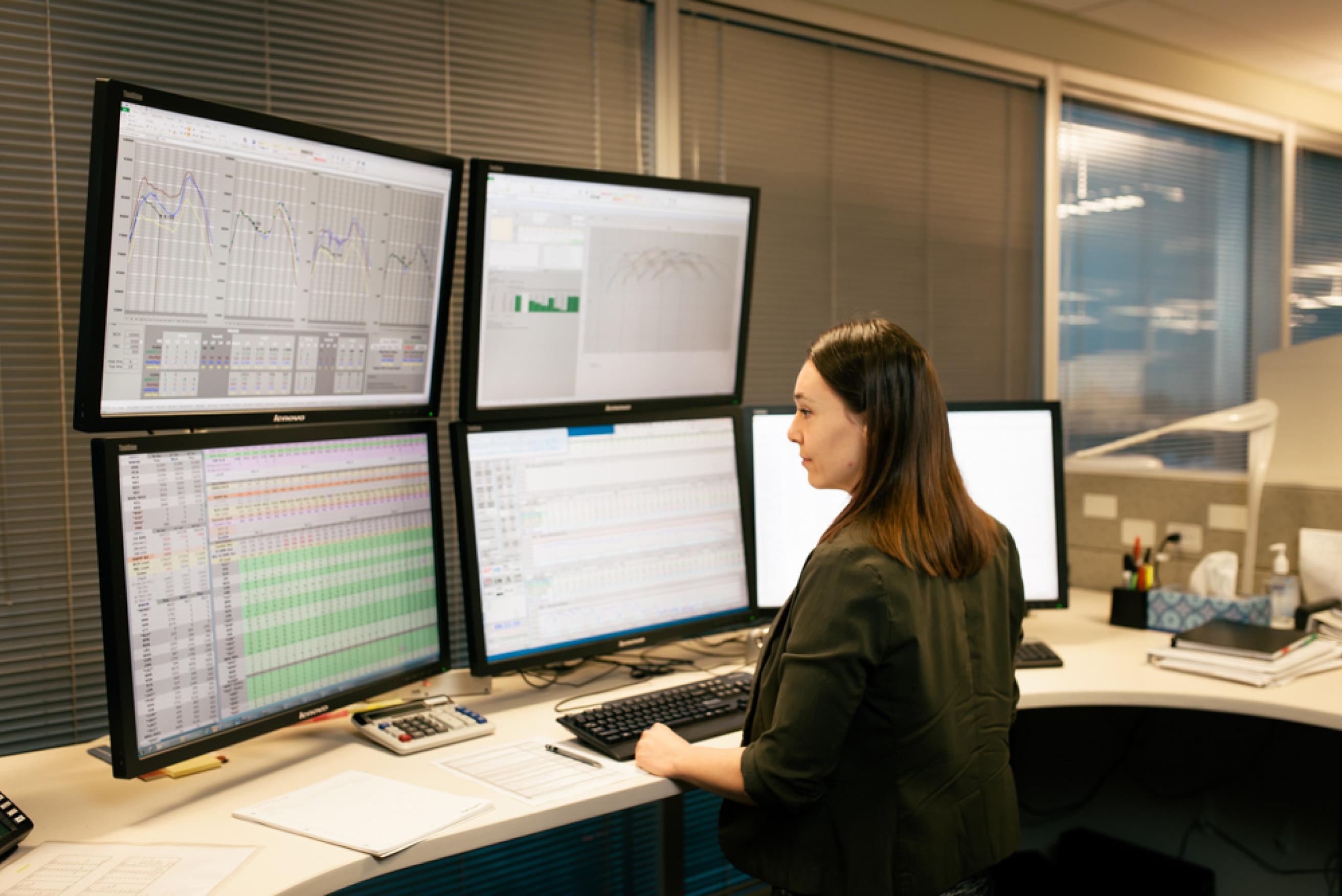 A woman standing at a desk with 5 monitors, faced away from the camera and working.