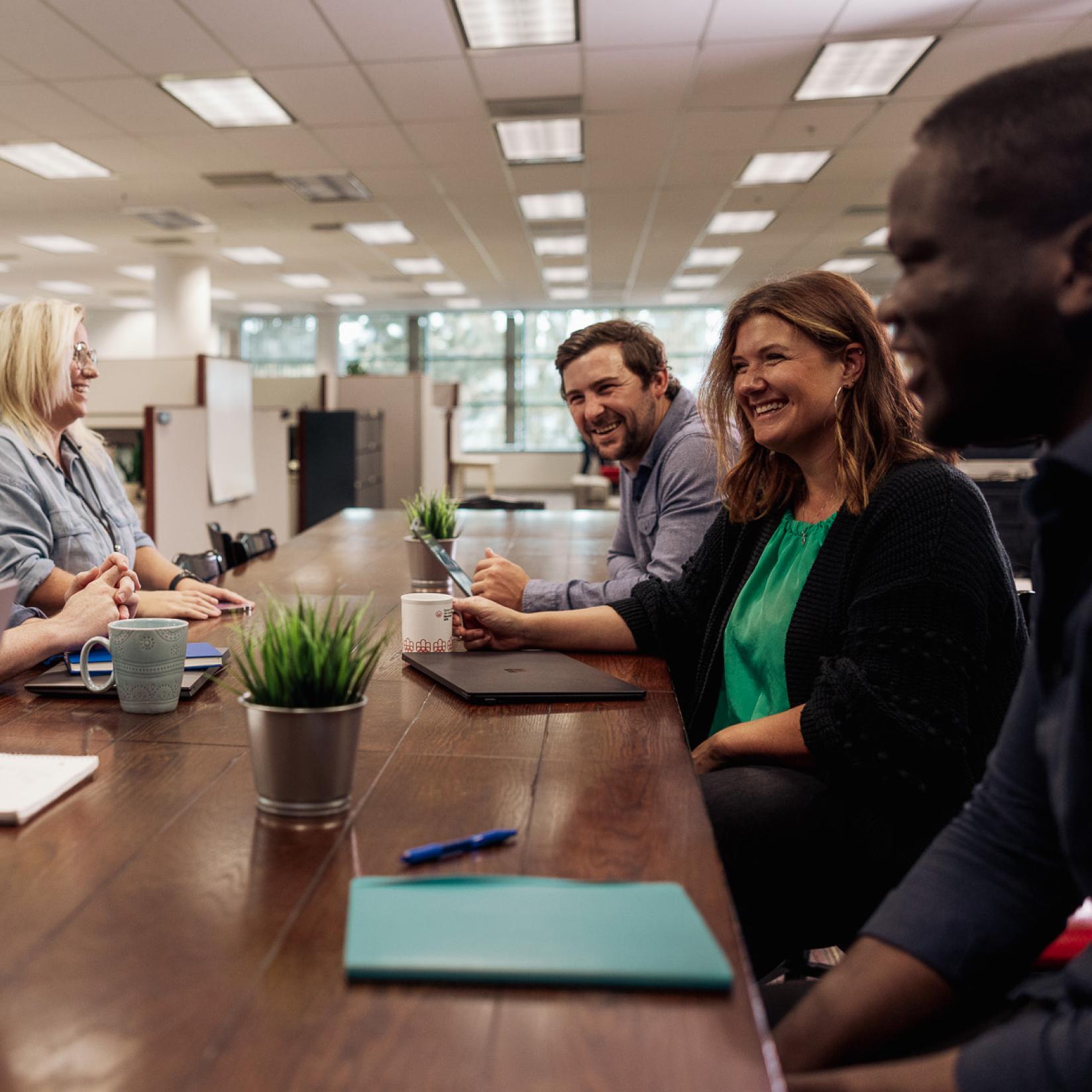 A group of people sitting at a desk, smiling at each other with papers on the table.