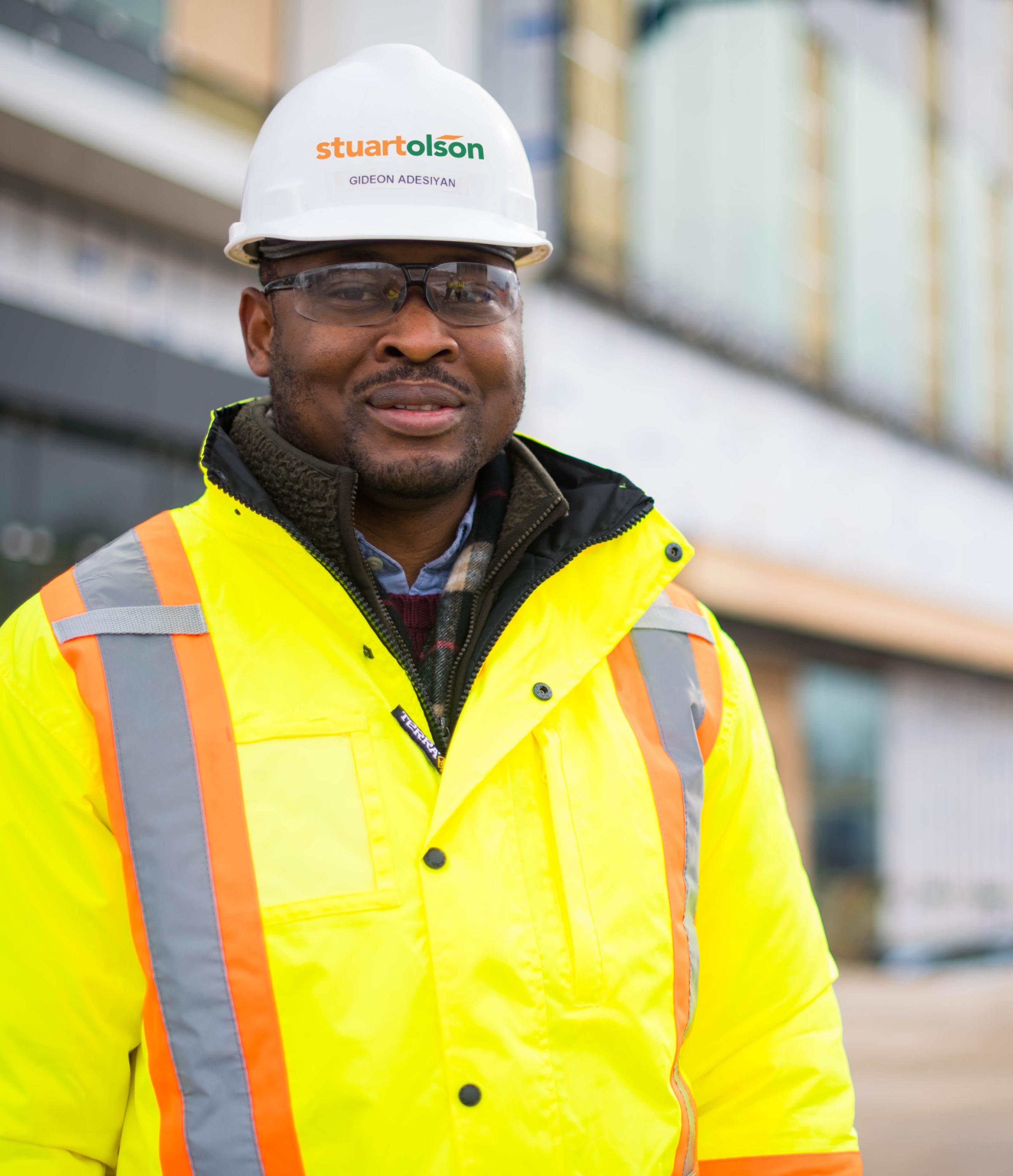A man standing outside of a building, smiling at the camera, wearing a safety jacket and a hard hat that says 'Stuart Olson'.