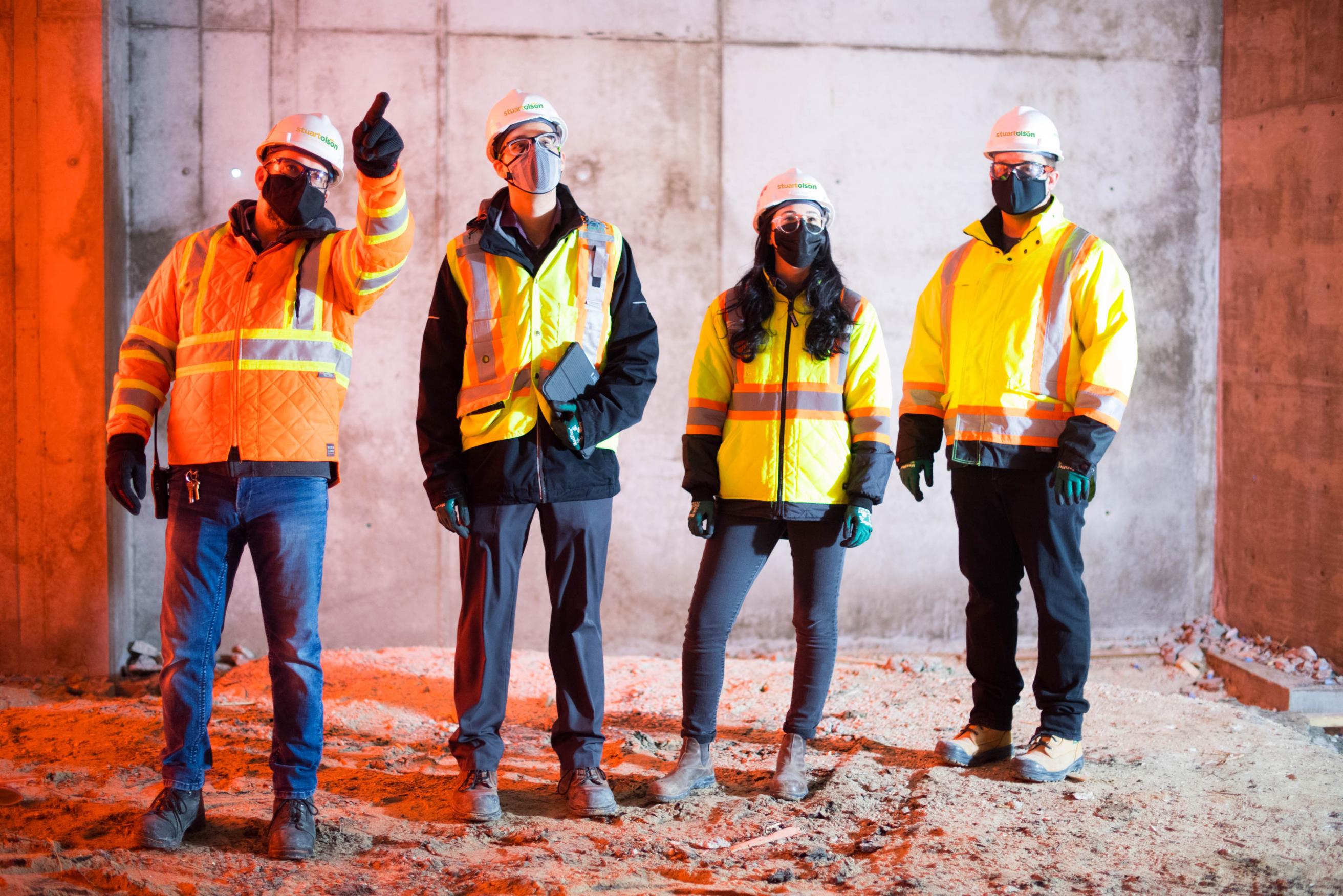 Four people in yellow safety jackets wearing hard hats inside of a unfinished building.