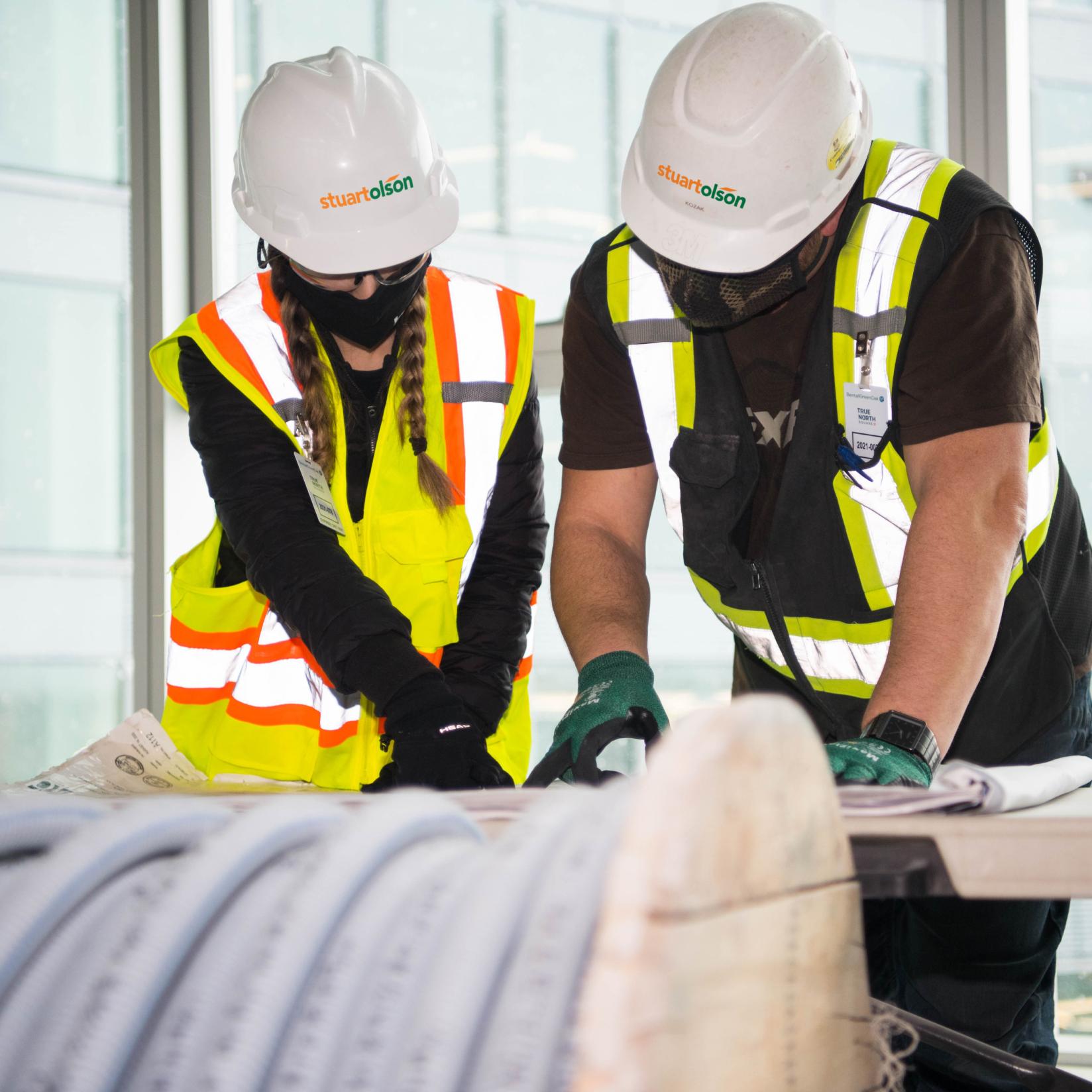 A woman and a man in safety vests and hard hats, leaning over a table to look at drawings on the table.