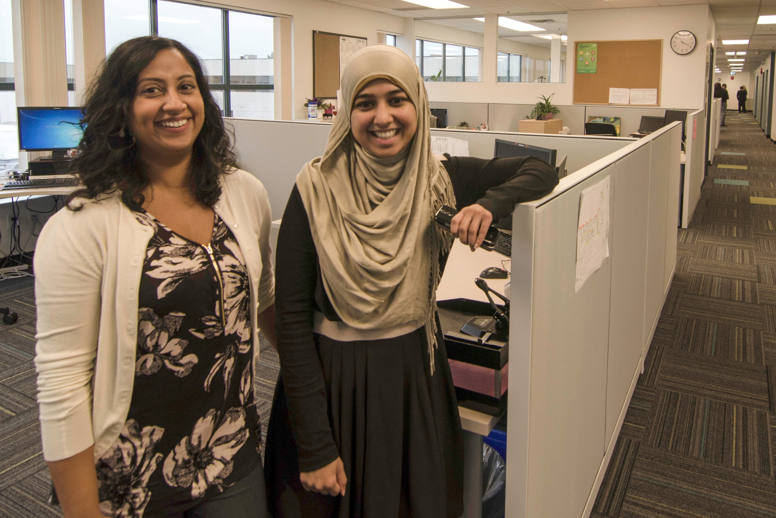 Two women standing by a desk, looking at the camera and smiling.