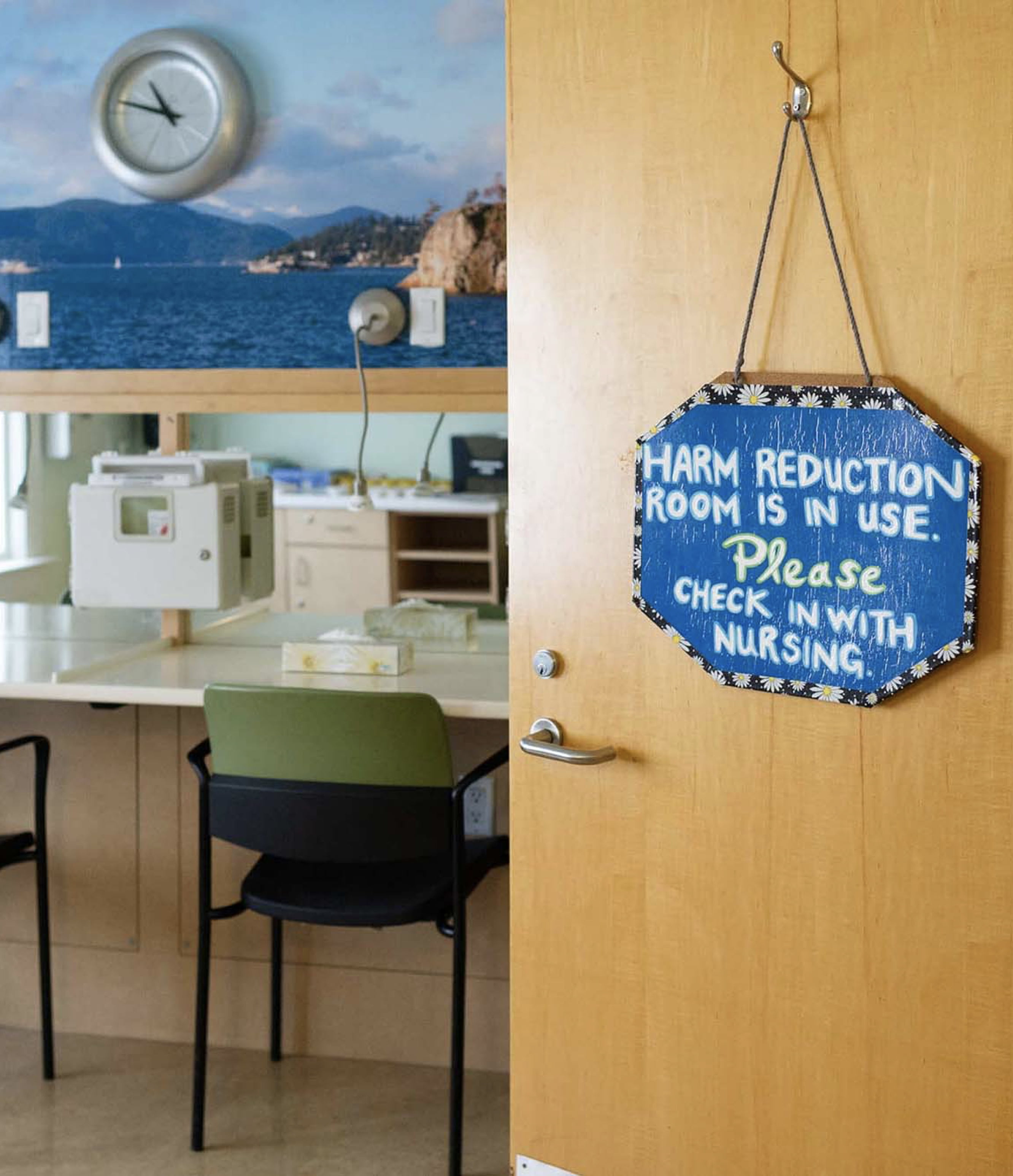 A door opening to a room with chairs with the sign 'Harm reduction room in use. Please check in with nursing' hanging from it.
