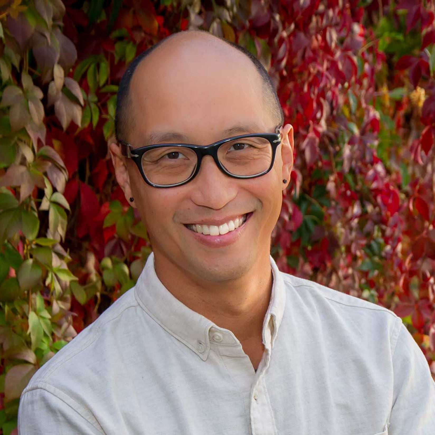 A man smiling at the camera in front of green, red and yellow leaves.