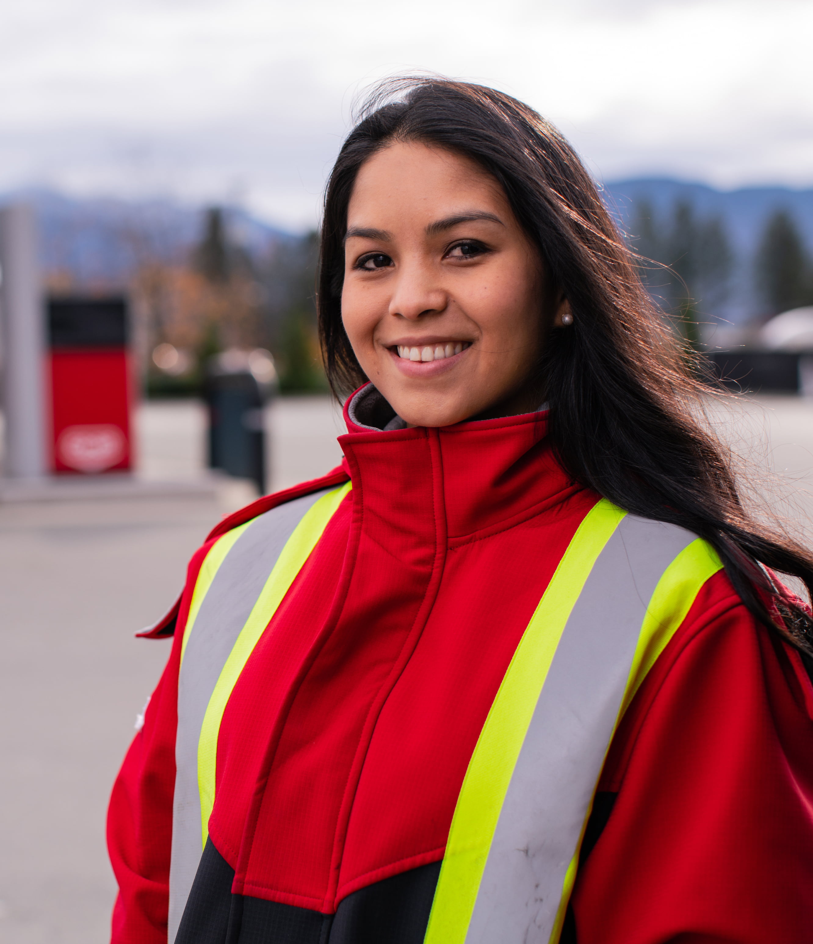 Woman outside of a Co-op gas station wearing a red jacket and safety vest, smiling at the camera.