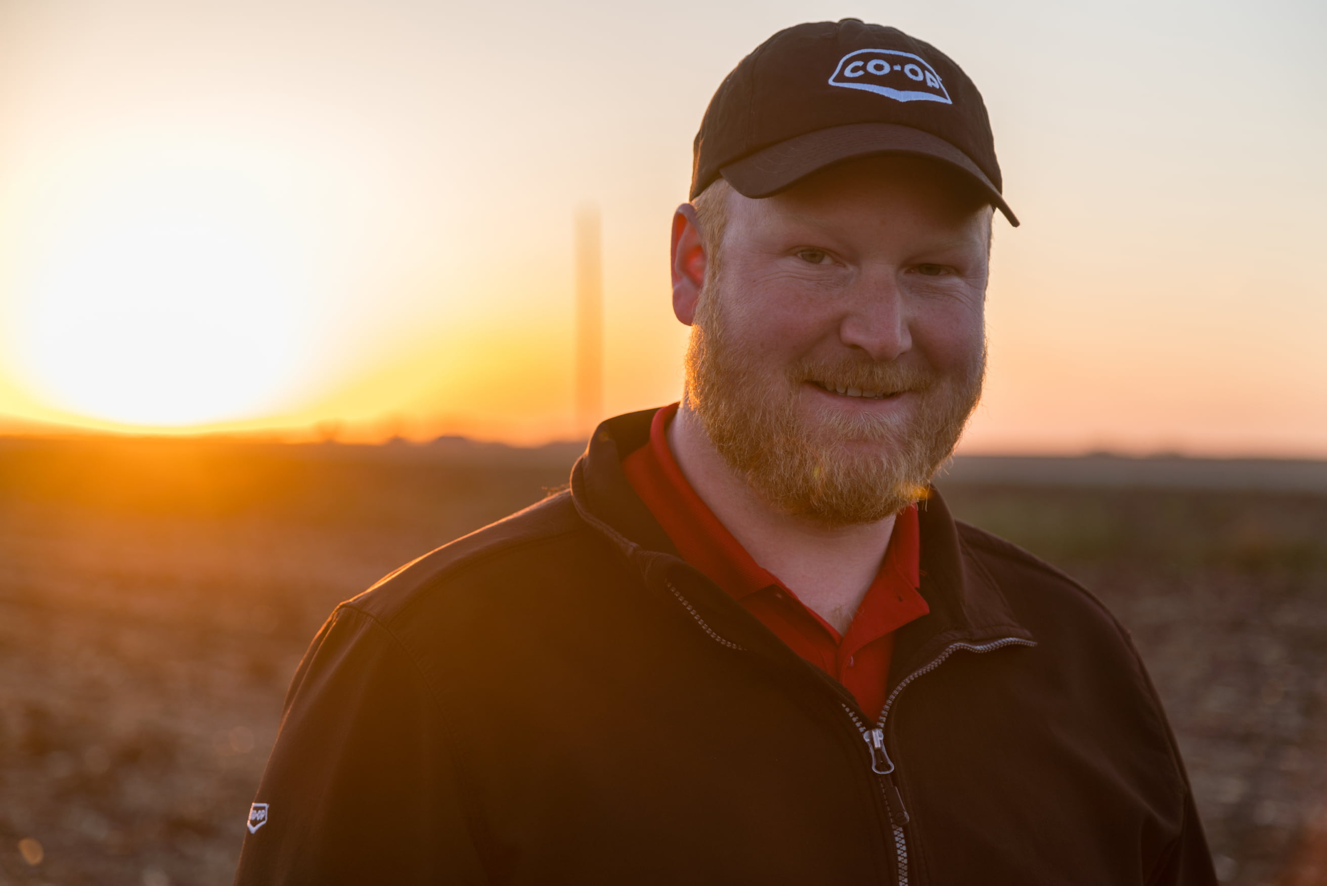 Man with a Co-op ball cap in a field, smiling at the camera with a sunset in the background.