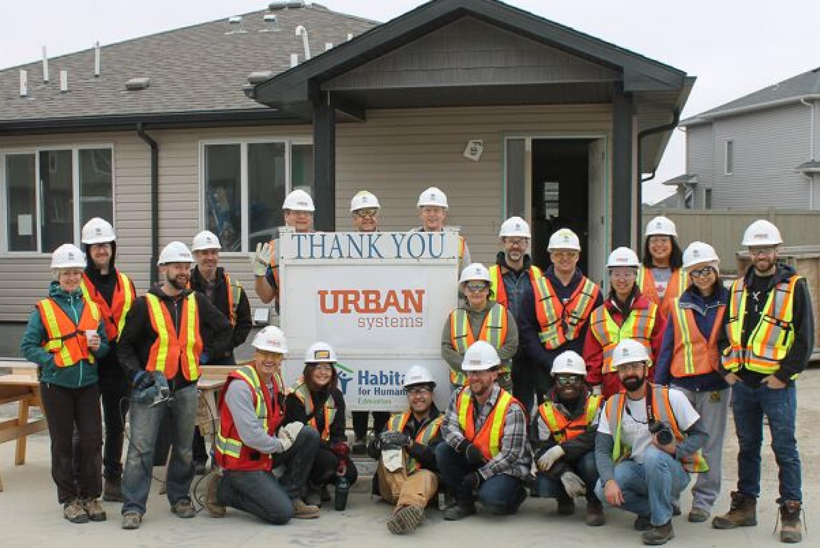 Group of Urban Systems construction workers standing outside of a house holding a sign.