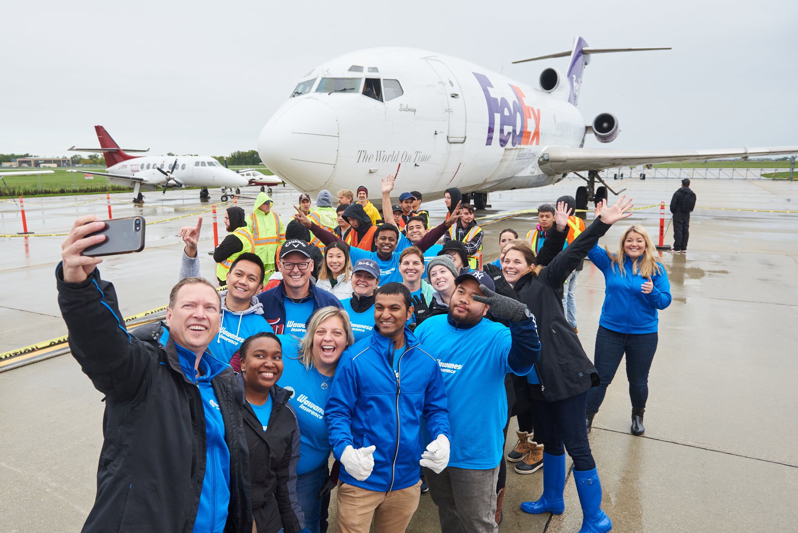 A group of Wawanesa employees in front of a FedEx airplane, taking a group selfie.